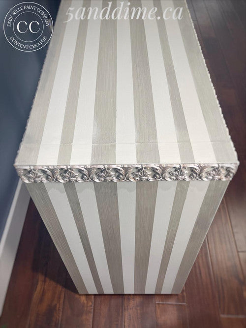 A striped rectangular chest with alternating light gray and white vertical stripes, decorative silver floral trim along the top edge, placed on a dark wooden floor next to a blue wall.