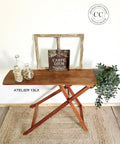 A wooden folding table holds decorative items, including a board with "Carpe Diem" text, two bottles with wood pieces and flower petals, and faux green foliage. A circular logo in the upper right corner reads "Dixie Belle Paint Company, Content Creator." Flooring is light wood.