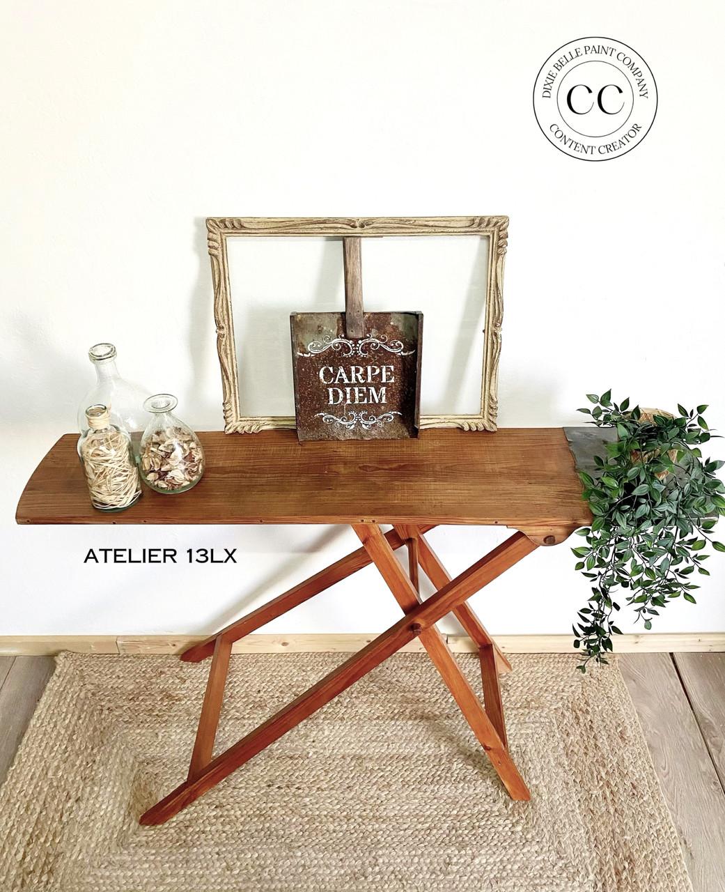 A wooden folding table holds decorative items, including a board with "Carpe Diem" text, two bottles with wood pieces and flower petals, and faux green foliage. A circular logo in the upper right corner reads "Dixie Belle Paint Company, Content Creator." Flooring is light wood.