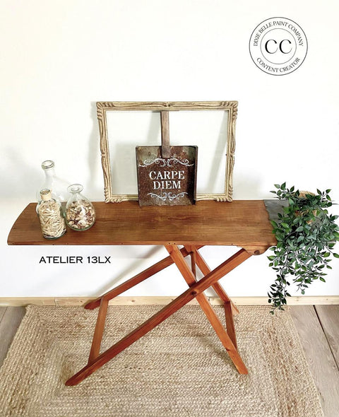A wooden folding table holds decorative items, including a board with "Carpe Diem" text, two bottles with wood pieces and flower petals, and faux green foliage. A circular logo in the upper right corner reads "Dixie Belle Paint Company, Content Creator." Flooring is light wood.
