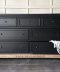 A black dresser with seven drawers, each featuring a round brass handle. The dresser has a light wooden base with short round legs. A textured grayish-blue cloth is draped over the right side. The wall behind is light-colored.