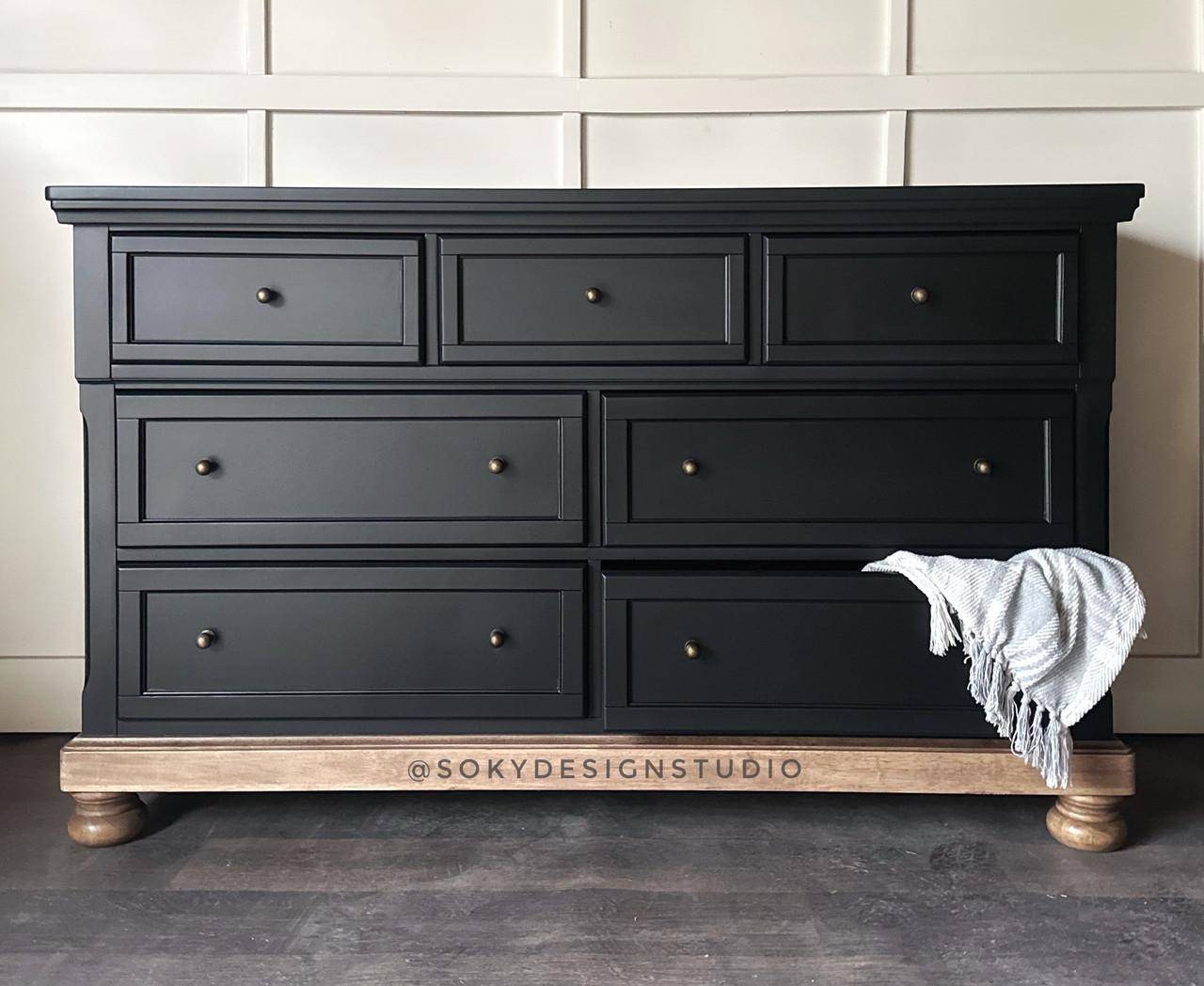 A black dresser with seven drawers, each featuring a round brass handle. The dresser has a light wooden base with short round legs. A textured grayish-blue cloth is draped over the right side. The wall behind is light-colored.