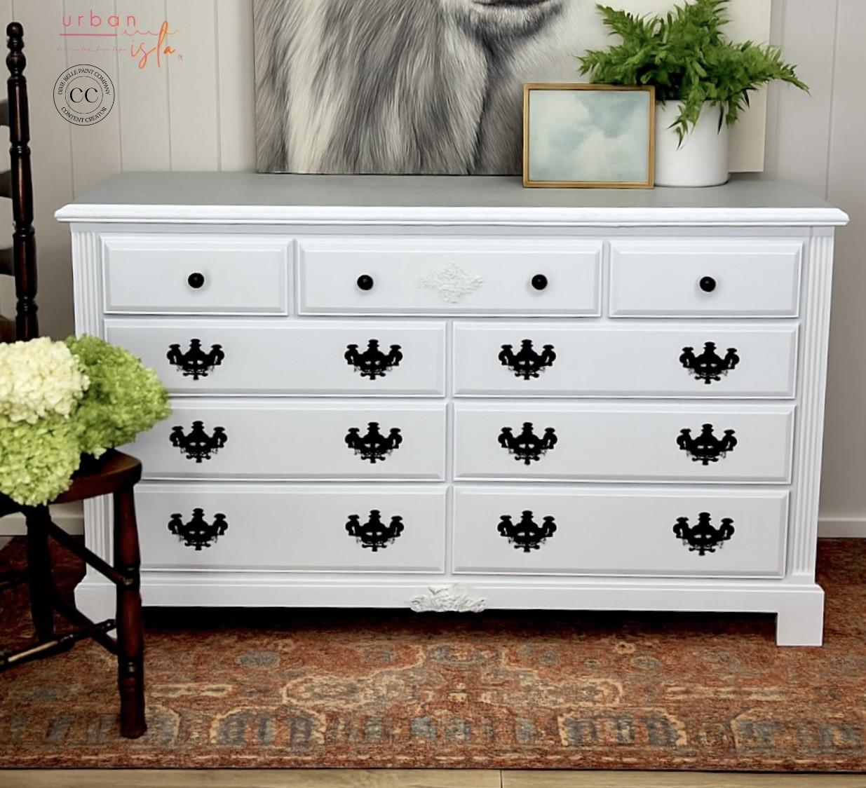 A white dresser with black handles stands on an ornate rug. On top are a framed abstract painting, a potted plant, and a green floral arrangement. A chair is partially visible to the left, and a black-and-white animal artwork is behind.