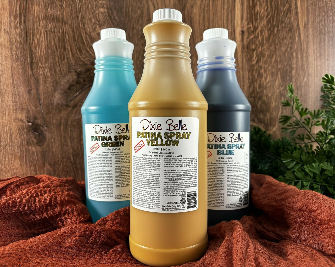Image of Yellow, Green, and Blue 32 oz. Patina Spray Refills. Staged on an orange blanket with faux greenery and faux wood behind it.