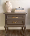 A taupe-colored vintage nightstand with ornate gold handles stands on a wooden floor. On top, there is a textured ceramic vase and two stacked books titled "ART AND LIFE IN AMERICA" and "MICHELANGELO.