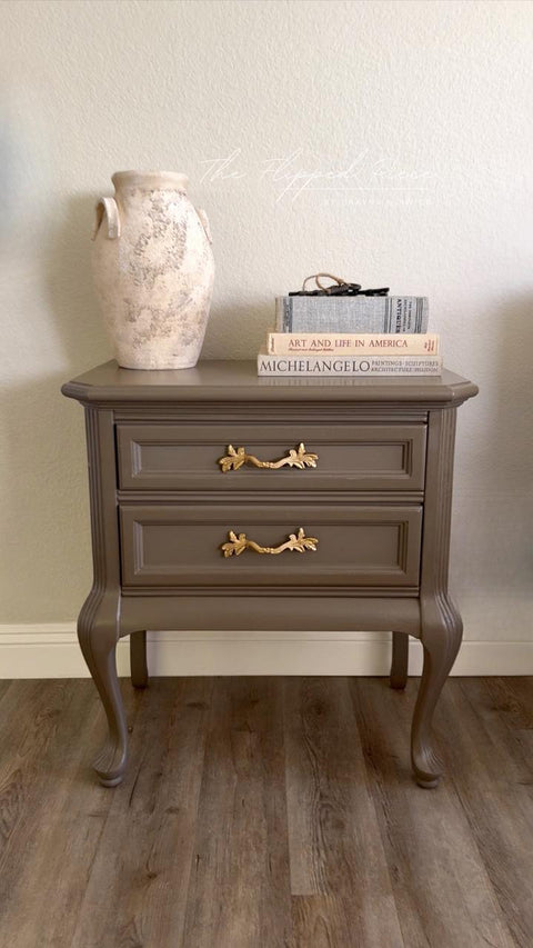 A taupe-colored vintage nightstand with ornate gold handles stands on a wooden floor. On top, there is a textured ceramic vase and two stacked books titled "ART AND LIFE IN AMERICA" and "MICHELANGELO.