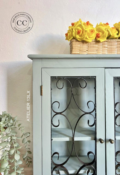 A light blue cabinet with ornate black metal doors stands against a white wall. On top, a wicker basket filled with yellow flowers is displayed. Green leaves cascade from the left side, and a round wall logo is visible.