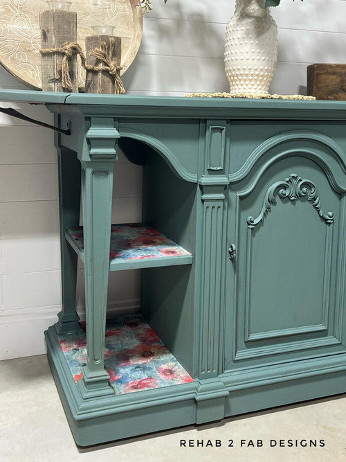 A teal wooden sideboard with ornate carvings on its doors and sides. The interior shelves are lined with colorful floral patterns. A vase with flowers and decorative books rest on top. Text reads "REHAB 2 FAB DESIGNS" on the bottom right corner.