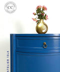 A blue dresser with a brass handle stands against a white wall. On top, there's a gold vase holding pink flowers. The wall has a logo with "CC" and "Content Creator," and the words "Atelier 13Lx" are vertically aligned beside the dresser.