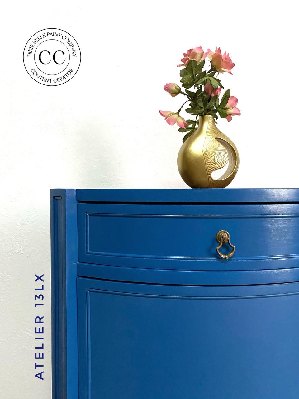 A blue dresser with a brass handle stands against a white wall. On top, there's a gold vase holding pink flowers. The wall has a logo with "CC" and "Content Creator," and the words "Atelier 13Lx" are vertically aligned beside the dresser.