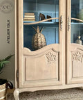 A rustic wooden cabinet with glass doors showcases decorative items, including stacked books, a woven basket, and small ornaments. The cabinet features ornate designs and is placed against a blue-painted interior background. Text reads "Au Naturel.