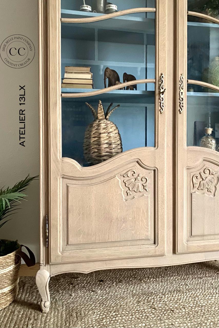 A rustic wooden cabinet with glass doors showcases decorative items, including stacked books, a woven basket, and small ornaments. The cabinet features ornate designs and is placed against a blue-painted interior background. Text reads "Au Naturel.