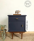 A dark blue wooden cabinet with brown legs and gold handles stands against a white wall. A small elephant figurine is placed on top. A green plant in a basket is on the left. The words "Atelier 13LX" are on the cabinet's side.