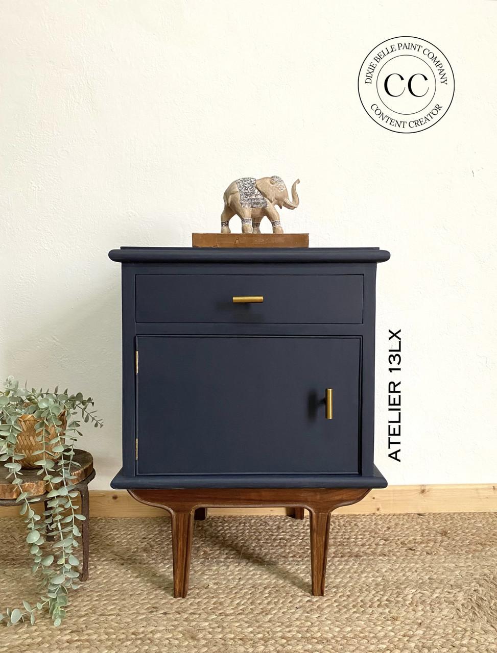 A dark blue wooden cabinet with brown legs and gold handles stands against a white wall. A small elephant figurine is placed on top. A green plant in a basket is on the left. The words "Atelier 13LX" are on the cabinet's side.