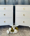 Two white ornate bedside tables with three embossed drawers each, featuring crystal knobs. They stand on a gray fluffy rug with white flowers in between. A textured blue wall is in the background.