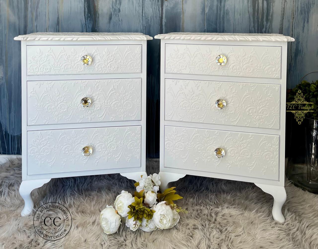 Two white ornate bedside tables with three embossed drawers each, featuring crystal knobs. They stand on a gray fluffy rug with white flowers in between. A textured blue wall is in the background.