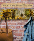 A coat rack with sunflower designs on a wooden board against a brick wall. The rack holds a brown handbag and a denim jacket.