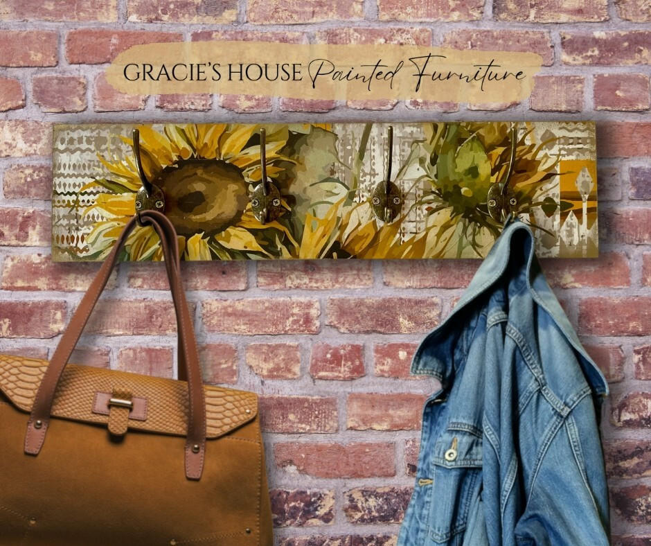 A coat rack with sunflower designs on a wooden board against a brick wall. The rack holds a brown handbag and a denim jacket.