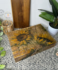Wooden box with intricate sunflower design, using the Indian Summer Decoupage Paper, rests on a textured surface among potted plants, candles, and rustic decor elements. The scene exudes a cozy, nature-inspired aesthetic with earthy colors and natural materials.