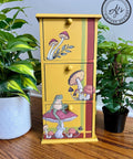 Yellow painted jewelry box with 3 drawers, with a green and red stripe on the right side and bottom. Mr. Toad's Transfer applied on the front of the drawers. Staged next to two pothos plants.