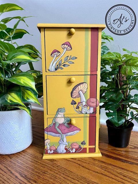 Yellow painted jewelry box with 3 drawers, with a green and red stripe on the right side and bottom. Mr. Toad's Transfer applied on the front of the drawers. Staged next to two pothos plants.