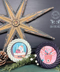 A festive display features two round framed holiday-themed paintings surrounded by pinecones and greenery. The left painting shows a snowy village scene, while the right painting depicts a reindeer with Christmas lights. Above them is a large rustic wooden star decoration.