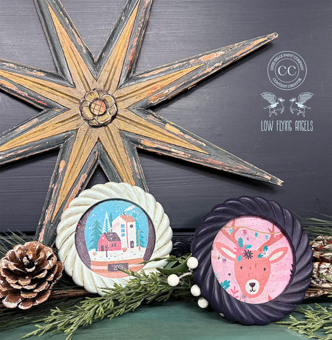 A festive display features two round framed holiday-themed paintings surrounded by pinecones and greenery. The left painting shows a snowy village scene, while the right painting depicts a reindeer with Christmas lights. Above them is a large rustic wooden star decoration.