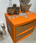 An orange Nightstand table with the Kaleidoscope Transfer applied to the top of the table and on the side. Staged with woven basket and glass jars on top of the piece.