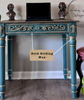 A teal table with ornate gold details stands on a wooden floor. A laptop is placed on top, along with two small bronze busts. A label pointing to the table reads "Gold Gilding Wax." A plant is visible to the side.