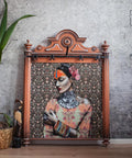 A framed portrait of a woman in traditional attire with colorful makeup, set against a floral background. The frame rests on the floor next to a small plant in a wicker pot and a decorative bird sculpture.