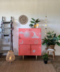 Pink painted desk with the Lani's Celestial Dream Transfer applied on the face of it and all 3 of the cabinets and drawers. Staged next to an old wooden ladder with flags hanging on it. A couple of potted plants and a candle on top. There are more plants on the floor and sitting on a stool next to the piece.