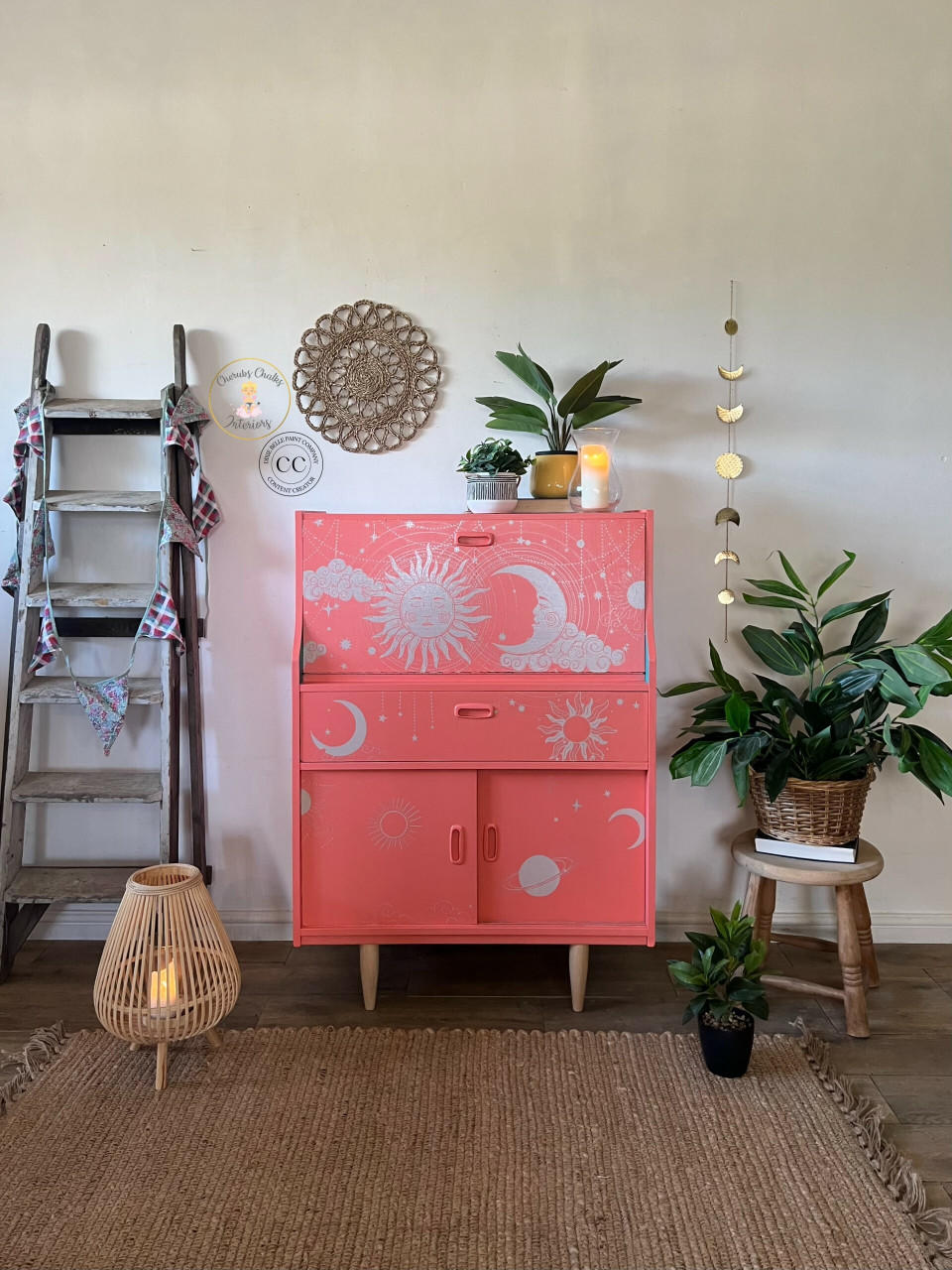 Pink painted desk with the Lani's Celestial Dream Transfer applied on the face of it and all 3 of the cabinets and drawers. Staged next to an old wooden ladder with flags hanging on it. A couple of potted plants and a candle on top. There are more plants on the floor and sitting on a stool next to the piece.