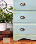 A close-up of a vintage dresser painted in a layered blue agave and pistachio finish with brass handles. A potted plant and two stacked books sit beside it on a wooden floor. A logo reads "AJ's Vintage Designs.