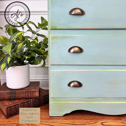 A close-up of a vintage dresser painted in a layered blue agave and pistachio finish with brass handles. A potted plant and two stacked books sit beside it on a wooden floor. A logo reads "AJ's Vintage Designs.