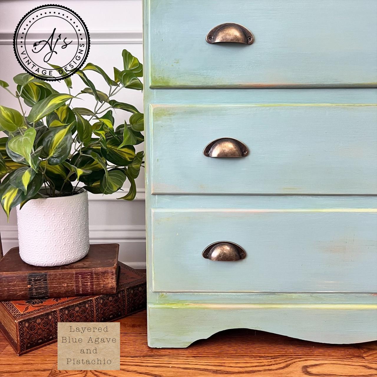 A close-up of a vintage dresser painted in a layered blue agave and pistachio finish with brass handles. A potted plant and two stacked books sit beside it on a wooden floor. A logo reads "AJ's Vintage Designs.