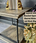 A rustic wooden table with two shelves. The top shelf holds books between decorative gold bookends. On the bottom shelf is a bouquet of dried flowers. The wall features a subtle floral pattern. Text reads: "Layered with Bleach Cypress, Charred Hickory, and Black Magic.