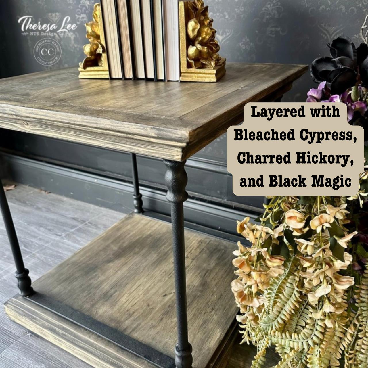 A rustic wooden table with two shelves. The top shelf holds books between decorative gold bookends. On the bottom shelf is a bouquet of dried flowers. The wall features a subtle floral pattern. Text reads: "Layered with Bleach Cypress, Charred Hickory, and Black Magic.