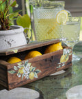 A rustic wooden box, with the Lemon Zest Transfer applied on front and top, filled with lemons on a glass table, with a pitcher of lemonade and a glass next to it. Natural outdoor setting with plants.