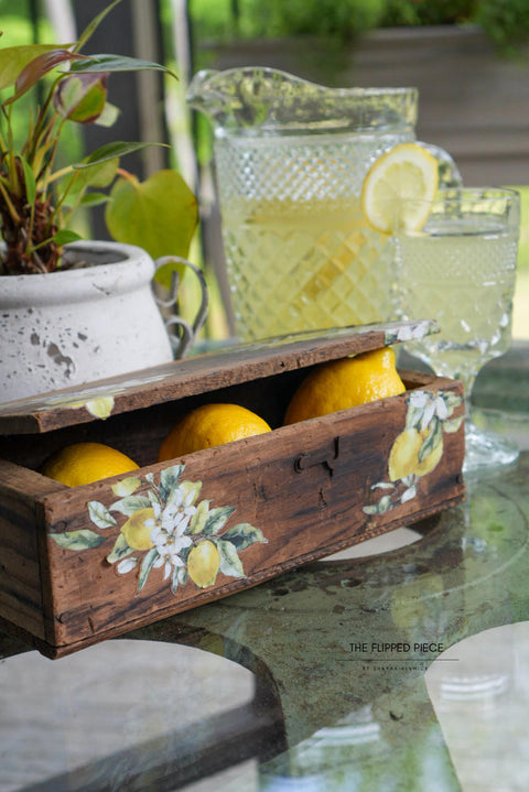 A rustic wooden box, with the Lemon Zest Transfer applied on front and top, filled with lemons on a glass table, with a pitcher of lemonade and a glass next to it. Natural outdoor setting with plants.