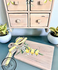 A wooden cutting board with the Lemon Zest Transfer applied, positioned in front of a wooden spice cabinet with the Lemon Zest Transfer applied on there also. Decorated with a yellow and burlap ribbon, placed near green plants and a blue wall.