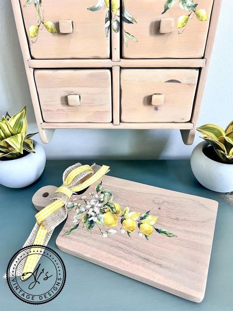 A wooden cutting board with the Lemon Zest Transfer applied, positioned in front of a wooden spice cabinet with the Lemon Zest Transfer applied on there also. Decorated with a yellow and burlap ribbon, placed near green plants and a blue wall.