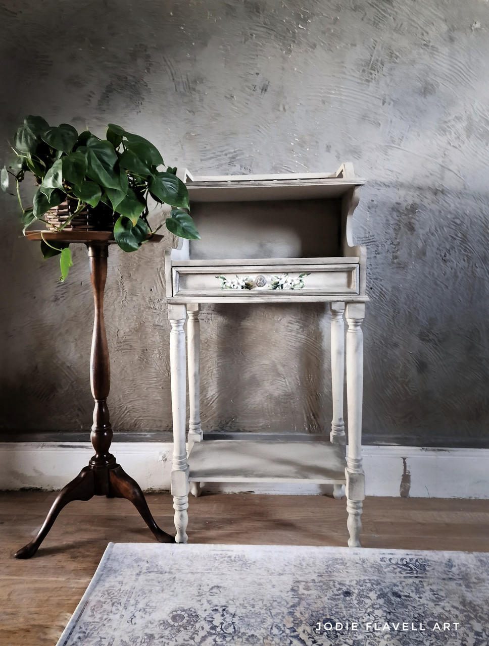 A vintage, white wooden console table, with the Lemon Zest Transfer applied on the drawer, stands against a textured grey wall, accompanied by a potted green plant on an ornate wooden stand and a decorative rug beneath.
