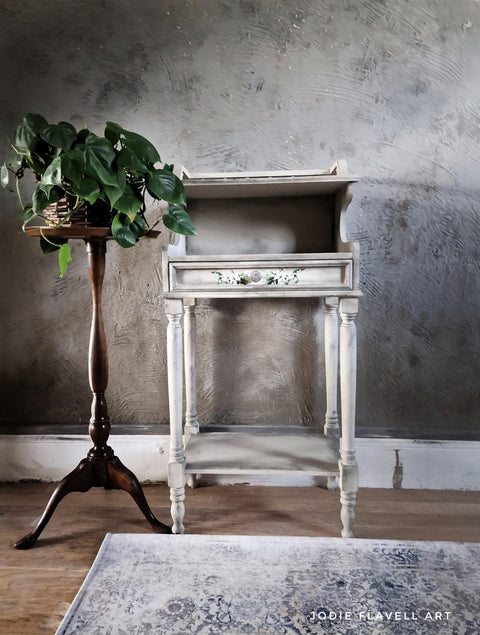 A vintage, white wooden console table, with the Lemon Zest Transfer applied on the drawer, stands against a textured grey wall, accompanied by a potted green plant on an ornate wooden stand and a decorative rug beneath.