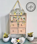 A vintage wooden spice cabinet with the Lemon Zest Transfer applied on the front and top, hung on a wall, flanked by two potted plants, with a bowl of seeds in the foreground.