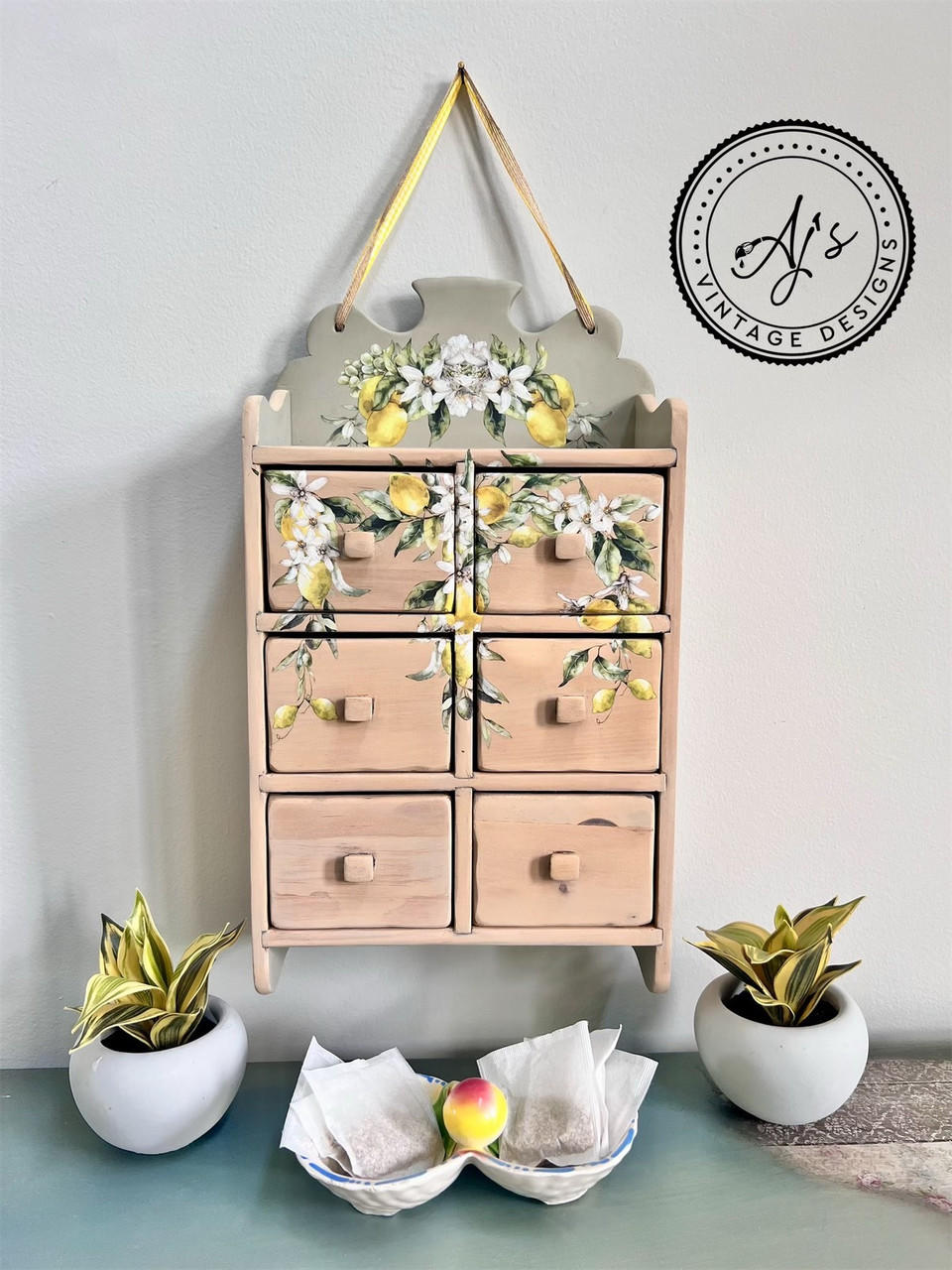 A vintage wooden spice cabinet with the Lemon Zest Transfer applied on the front and top, hung on a wall, flanked by two potted plants, with a bowl of seeds in the foreground.