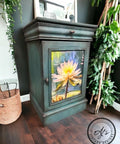 A dark green painted wooden cabinet with Lily Pads Decoupage Paper applied on its door, positioned in a corner next to a basket and artificial greenery in a cozy room setting.