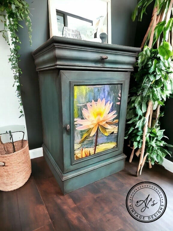 A dark green painted wooden cabinet with Lily Pads Decoupage Paper applied on its door, positioned in a corner next to a basket and artificial greenery in a cozy room setting.