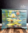 A painted wooden dresser with vibrant water lilies and lily pads on a pond. The dresser features four drawers, each adorned with metal handles and the text "Tracey's Fancy" in the upper right corner of the black background wall.