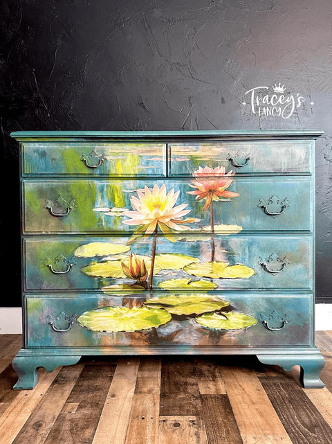 A painted wooden dresser with vibrant water lilies and lily pads on a pond. The dresser features four drawers, each adorned with metal handles and the text "Tracey's Fancy" in the upper right corner of the black background wall.