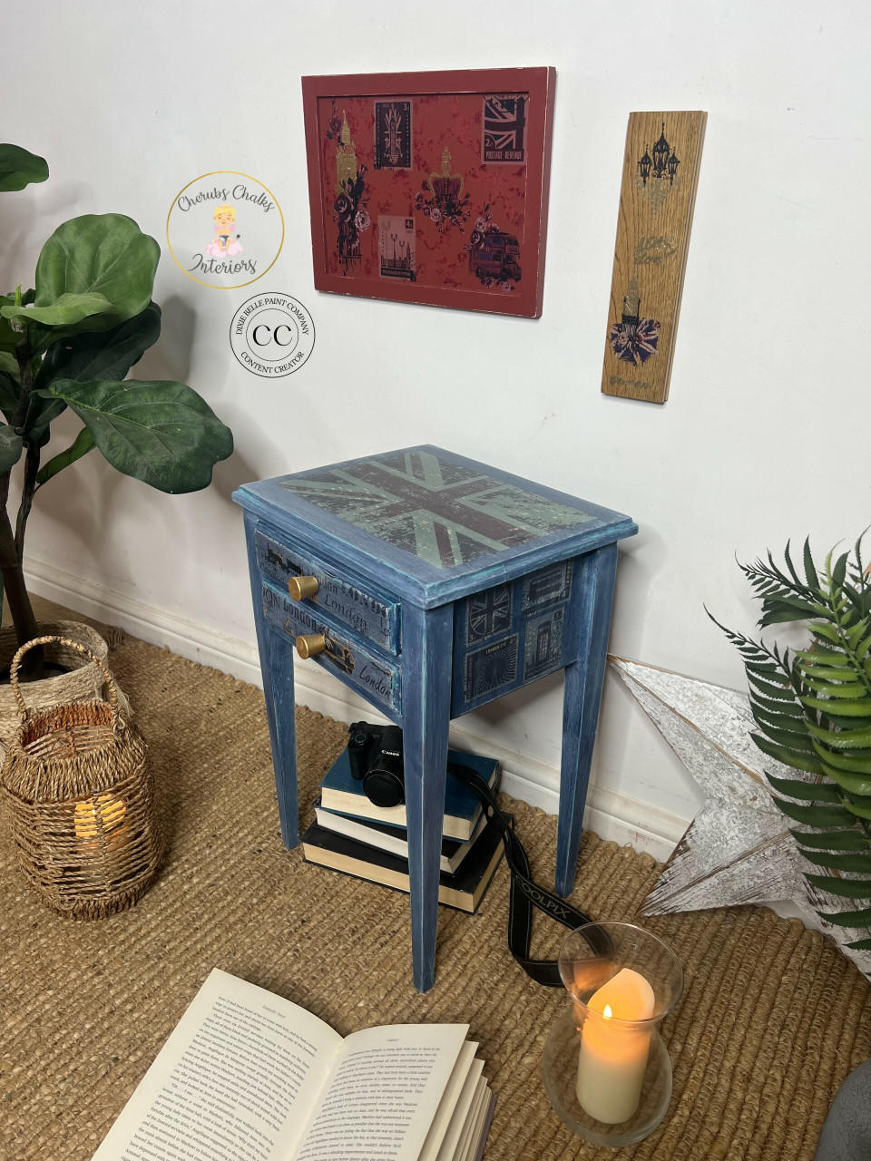 Blue painted and distressed end table with large gold knobs on the 2 drawers. The London Vibe Transfer has been applied to the top, front, and side of the piece. Staged with a stack of books underneath it with a camera on top. The London Vibe Transfer has also been applied to some decorative pieces of wood that are hung on the wall.
